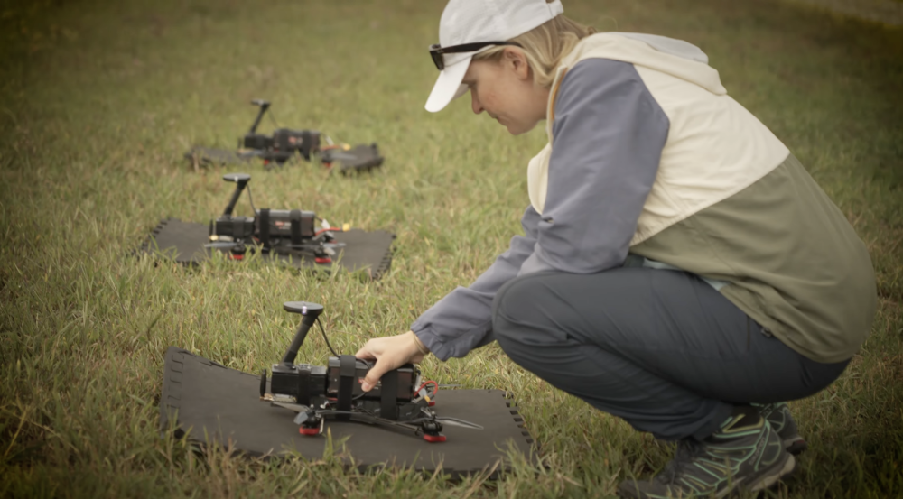 A staff member crouches beside three drones lying on the grass.