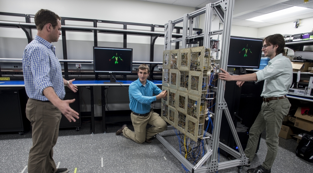 Researchers test the prototype standoff microwave imaging system. The antennas emit radio signals that reflect off the person standing in front of the array; the system processes the reflections to create the image on the monitors in the background.  