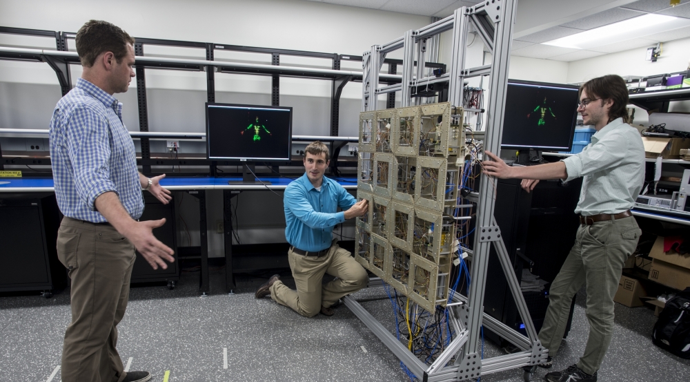 Researchers test the prototype standoff microwave imaging system. The antennas emit radio signals that reflect off the person standing in front of the array; the system processes the reflections to create the image on the monitors in the background.