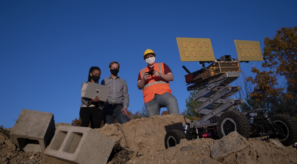 three people stand on a big dirt pile, outside, with blue sky in the background. One researcher is holding a laptop, which another looks at. The third person is holding a remote, controlling a robotic vehicle also on the dirt pile. 