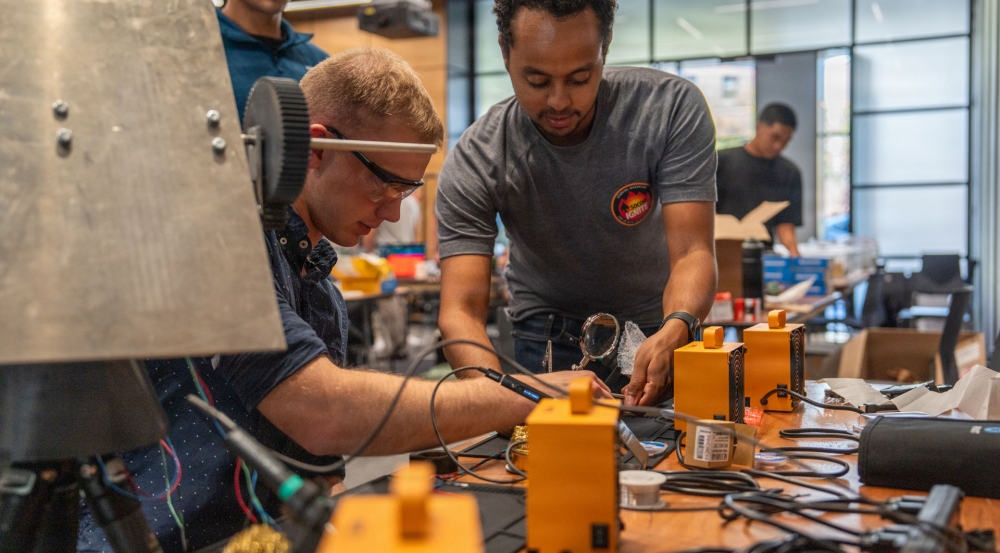 A technical mentor works alongside a student in a classroom, adjusting technical equipment on a table.