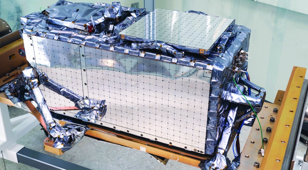One of the QZSS payloads is shown close up, sitting on a table. It is a shiny, silver-colored rectangular box with various protruding attachments.