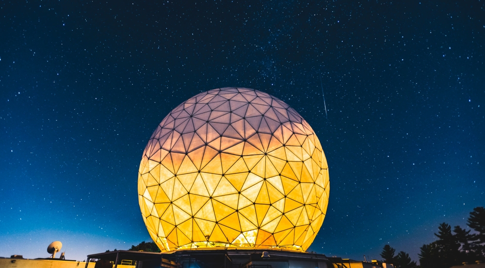 A large radar dome is brightly lit from the inside, shown in the foreground of a night sky full of stars.  