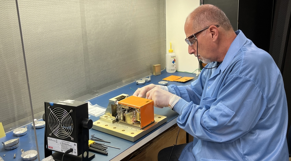 A technician sits at a bench working on a small metal payload. 