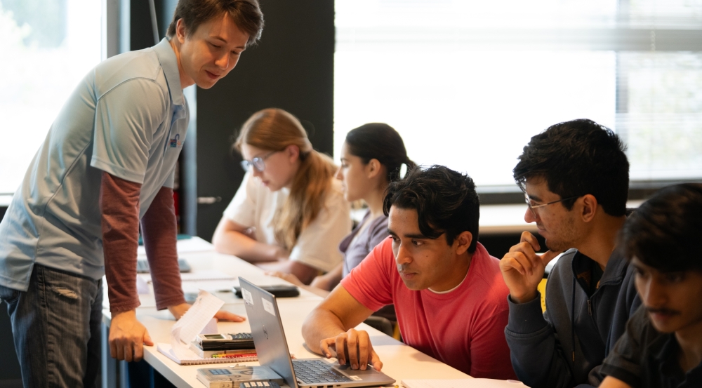 An instructor stands on one side of a classroom table in front of five students practicing advanced math for cybersecurity applications.   