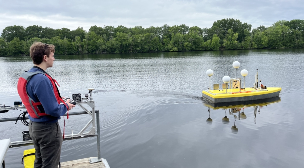 A researcher, operating a remote controller in his hands, stands on a dock while looking over at a small vessel (approximately 6 by 3 feet) containing sonar and antenna equipment on a river. 