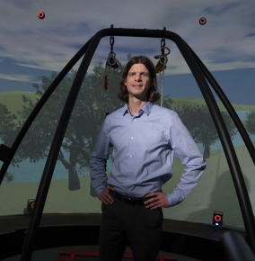A photo of Brian Baum standing on a treadmill with support beam surrounding him. In the background is a screen showing a cloudy sky and trees. He is standing is a virtual reality dome.