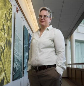 Molly Crane poses for a photo, smiling with her hands in her pockets, in a hallway. To her right is a wall with photos on it, one orange, then yellow, then blue. To her left is an indoor balcony.