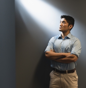 Yutai Zhou stands with his arms crossed, against a blue-grey wall. He looks up at light coming through a window.