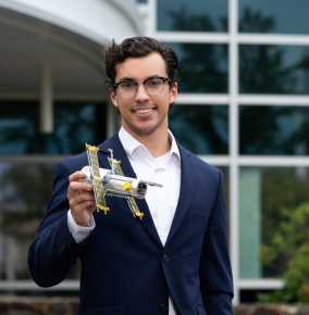 Philip Lopez stands at the front entrance of the Laboratory holding a small model satellite