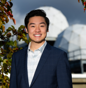 Nicholas Leung smiles while standing in front of two radomes and some colorful leaves on a sunny day. 