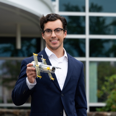 Philip Lopez stands at the front entrance of the Laboratory holding a small model satellite