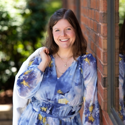 A photo of Theresa outside. She learning against a brick building and wearing a blue floral dress
