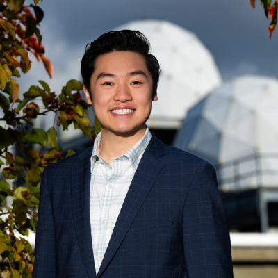 Nicholas Leung smiles while standing in front of two radomes and some colorful leaves on a sunny day. 