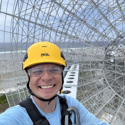 Andrew Dahir, wearing a yellow helmet, stands in front of a radar with the ocean in the backdrop.