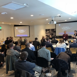 About thirty people sit in a classroom setting, with chairs facing a panel at front and projectors with a slide reading "Resilience in Complex Systems"