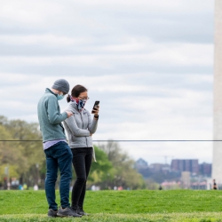 A photo of two people standing together wearing face masks, with the washington monument in the background. 