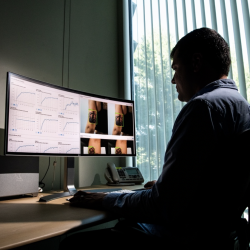 A man sits at an office desk looking at a large monitor. the left hand side shows data graphs, the right side shows two images of the back of a woman's arm with a crown tattoo, and a green box around the tattoo.