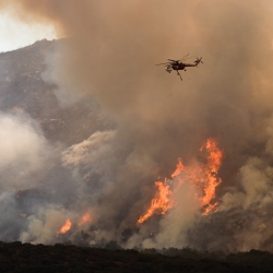 A helicopter flies closely over a wildfire on a mountain. You can see orange flames amongst a large cloud of white smoke. 