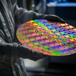 A person with a protective gown and gloves holds a large wafer of integrated circuits. the wafer is vividly multicolored.