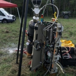 A photo of a tactical hydrogen generatation system. Setup outside on grass, the system is compose of a black tank, attached to a metal trunk containing wiring and hardware. 