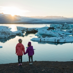 Two young girls hold hands while looking over glaciers and water. 
