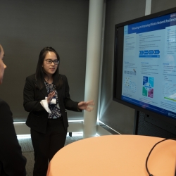 A researcher stands and points to screen showing her research poster. She is talking about the research with two people looking at the screen.
