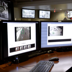 Two desktop monitors run software for forensic video analysis, with two additional monitors in the background capturing video surveillance from different angles. 