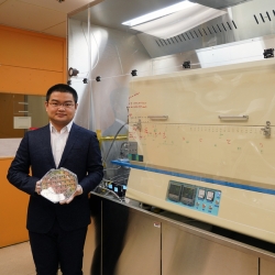 A researcher holds a wafer, while standing in front of a material deposition machine.