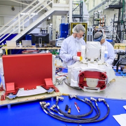 Three people examine an optical communications payload at Kennedy Space Center.