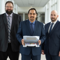 Three men stand side by side, with the person in the middle holding a box containing a software package.