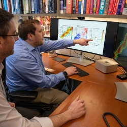 two scientists sit at a desk in front of two computer screens; the screens show radar images of tornadoes.