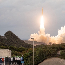 Spectators on a platform watch as the Quasi-Zenith Satellite System-Hosted Payload lifts off in the distance over a forest. 