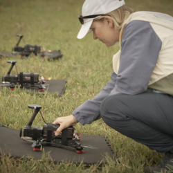 A staff member crouches beside three drones lying on the grass.