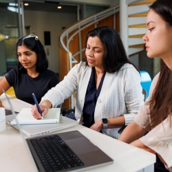Three people sit at a desk with laptops and a notepad.