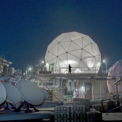 Four satellite dishes and two radomes, each covering satellite communications antennas, are shown on the roof of Lincoln Laboratory among other equipment.