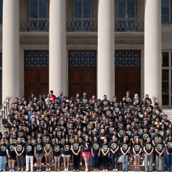 BWSI students wear matching black shirts with the BWSI logo and stand on steps outside of an MIT building with columns in the background. 
