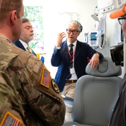 ARPA-H program manager Bon Ku faces the camera while talking to service members who are in the foreground. Part of the PSI vehicle's patient chair setup is visible next to Ku. 