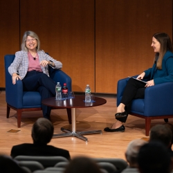 Sally Kornbluth and Melissa Choi sit in blue chairs on a wooden stage in Lincoln Laboratory's auditorium. 