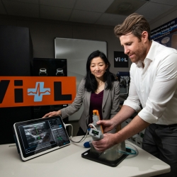 Two researchers standing at a table inside of a lab showing the AI-Guide handheld device and an ultrasound display.