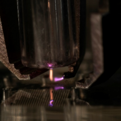A zoomed-in view of a metal nozzle shooting a stream of orange onto a metal surface, and a nozzle shooting a stream of purple at the orange stream of material. 