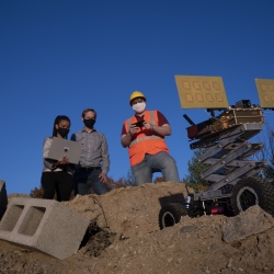 three people stand on a big dirt pile, outside, with blue sky in the background. One researcher is holding a laptop, which another looks at. The third person is holding a remote, controlling a robotic vehicle also on the dirt pile. 