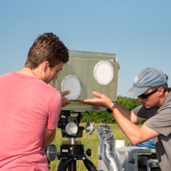 Two men adjust a metal antenna unit, approximately 12 in by 18 in, on a tripod.