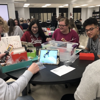 A group of five high school students sit at a table, smiling, working together to assemble a robot out of what look to be lego-like parts. 