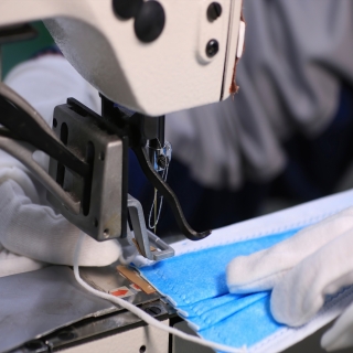 An image of a person (only their hands are shown) using a sewing machine to sew a blue surgical mask.