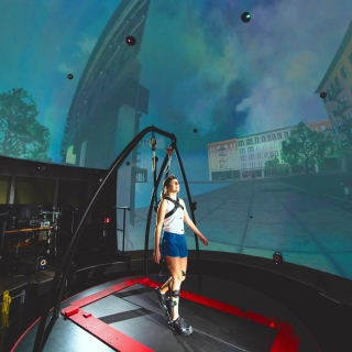 A woman, attached to a harness, stands on a treadmill, and is surrounded by a spherical screen displaying a city scene.