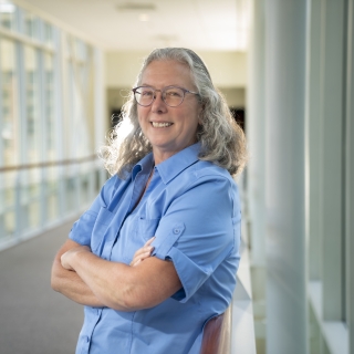 Mary Ellen Zurko poses for a photo in a sunny, glass hallway. She's wearing a light blue shirt and her arms are crossed in front of her.