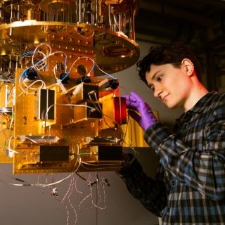 Alex Green stands at a dilution refrigerator, a gold/metal contraption. 