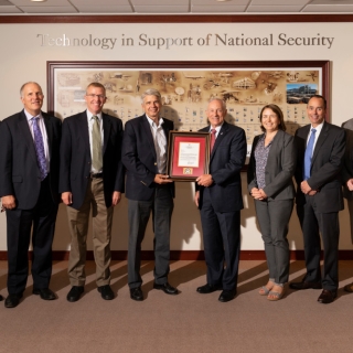 A photograph of eight individuals, with the two in the center holding a plaque in front of a wall that says "Technology in Support of National Security."