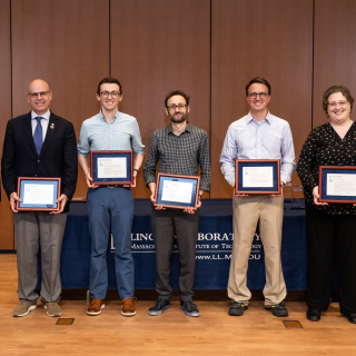 A group of six people pose a group photo on a stage. Five of the people hold award plaques.